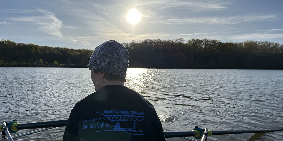 Back of a rower at sunrise wearing a Mid-Hudson Rowing long-sleeve tee. 